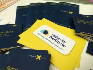Blaue Informationsbroschüren der Opferhilfe-Berlin umrahmen gelbe Informationskarten des Online-Portals Hilfe-in-Berlin.de