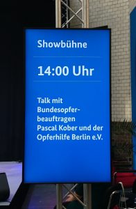 Zu sehen ist ein rechteckiger, blauer Screen, der einen Programmpunkt ankündigt: Showbühne, 14 Uhr, Talk mit dem Bundesopferbeauftragten Pascal Kober und der Opferhilfe Berlin e.V.