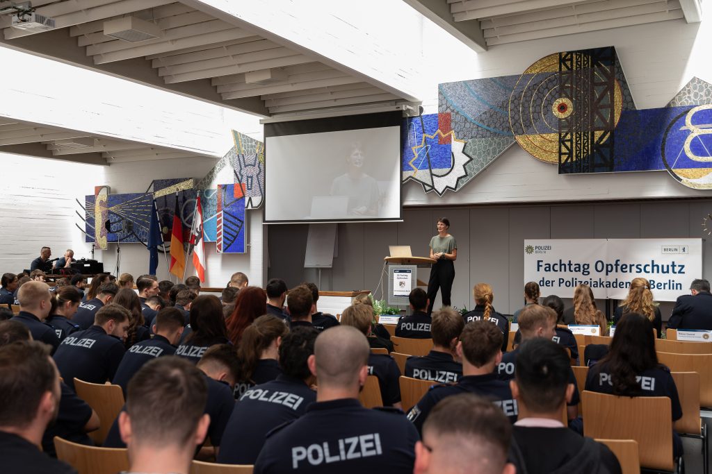 Das Bild zeigt einen Vortragssaal, in dem viele Menschen in Polizeiuniform sitzen. Auf der Bühne steht eine Referentin von proaktiv.